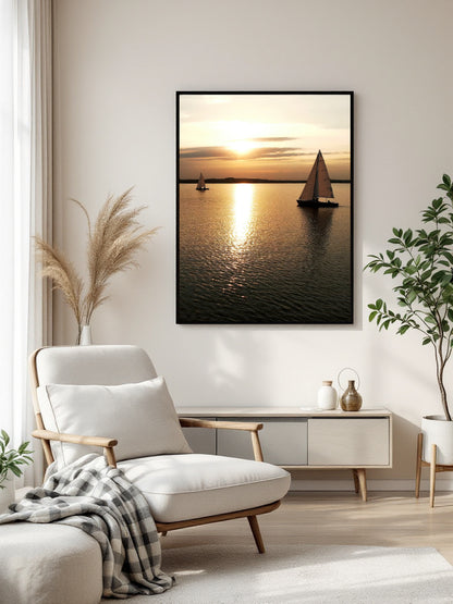 A framed vertical canvas print of a sunset over the water with sailboats, displayed on a light beige wall in a modern living room setting with a white armchair, console table, and potted plants.