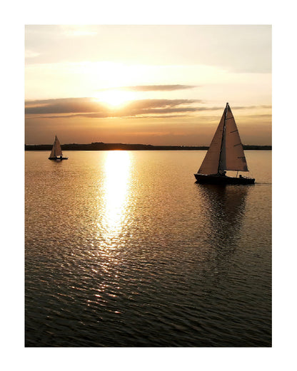 A sunset seascape print featuring two sailboats on calm water with the sun low on the horizon, casting a bright golden reflection across the surface.