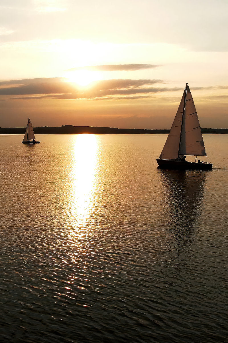 Two sailboats on calm water at sunset, with the sun low over the horizon and golden light reflecting across the water surface.