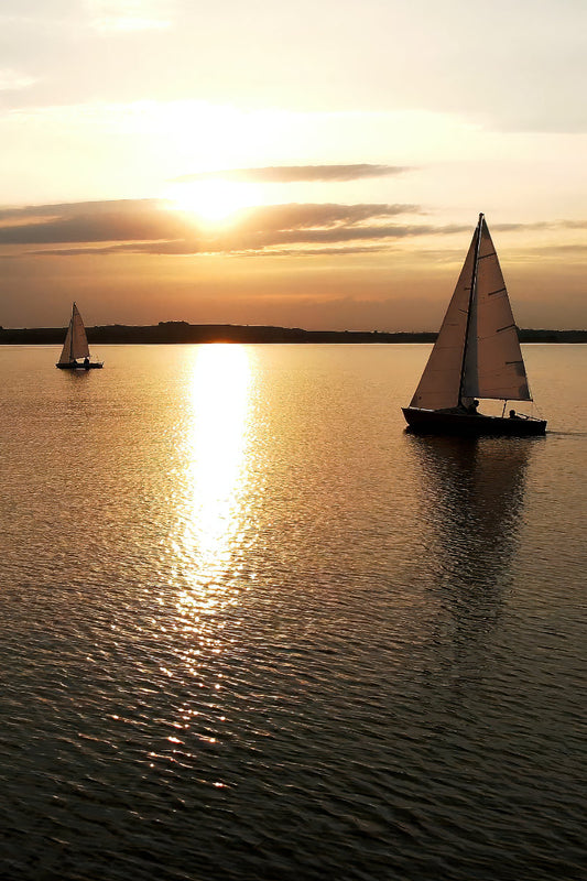 Two sailboats on calm water at sunset, with the sun low over the horizon and golden light reflecting across the water surface.