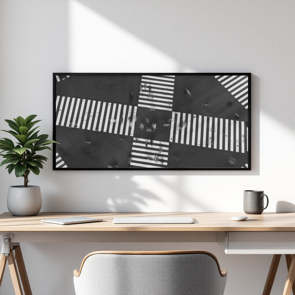 A framed black and white abstract geometric wall art print hangs on a white wall above a light wood desk with a laptop, mouse, mug, and potted plant.