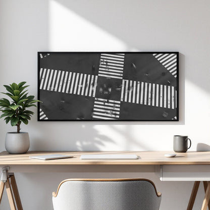 A framed black and white abstract geometric wall art print hangs on a white wall above a light wood desk with a laptop, mouse, mug, and potted plant.