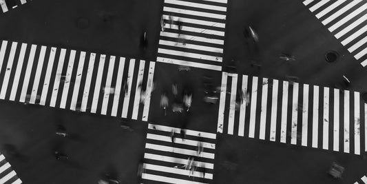 Black and white aerial photograph of a city intersection with striped crosswalks and blurred figures and vehicles moving through the crossing.