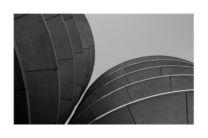 Black and white architectural photograph of two curved dome-like structures with linear paneling, presented with a white border.