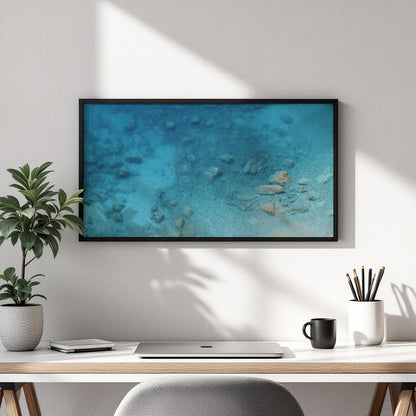 A framed wall art print showing an underwater ocean scene in shades of blue and turquoise, hung on a white wall above a desk with a plant, laptop, mug, and pencils.