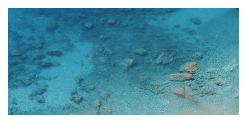 Underwater ocean scene with clear turquoise-blue water, shallow sandy seabed, and scattered rocks.
