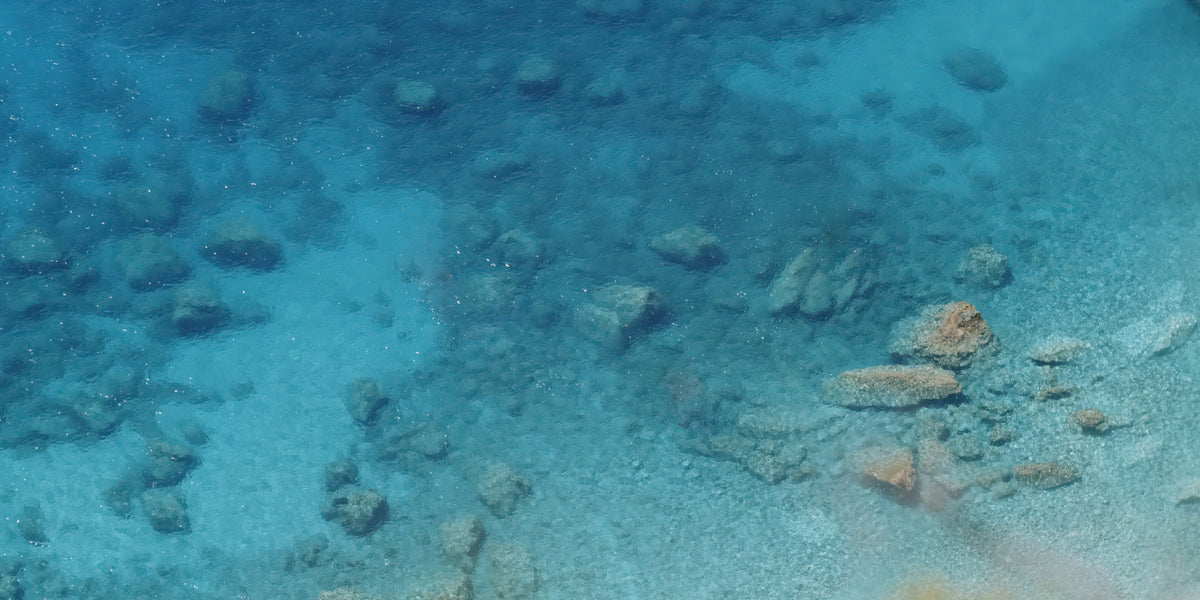 Underwater clear blue water showing a rocky and sandy seabed with small stones and coral-like formations.