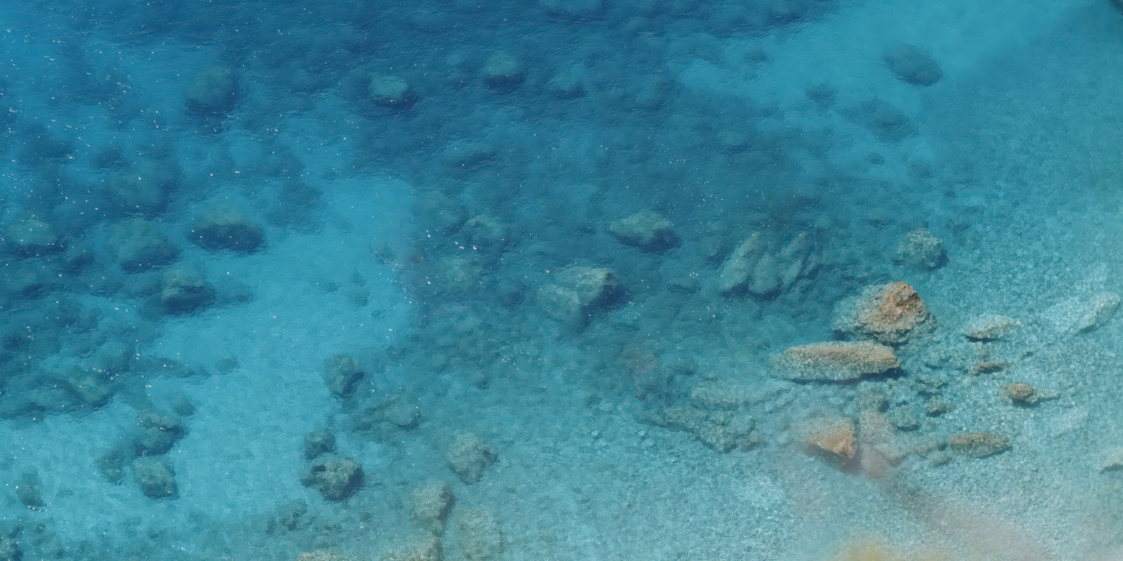 Underwater clear blue water showing a rocky and sandy seabed with small stones and coral-like formations.