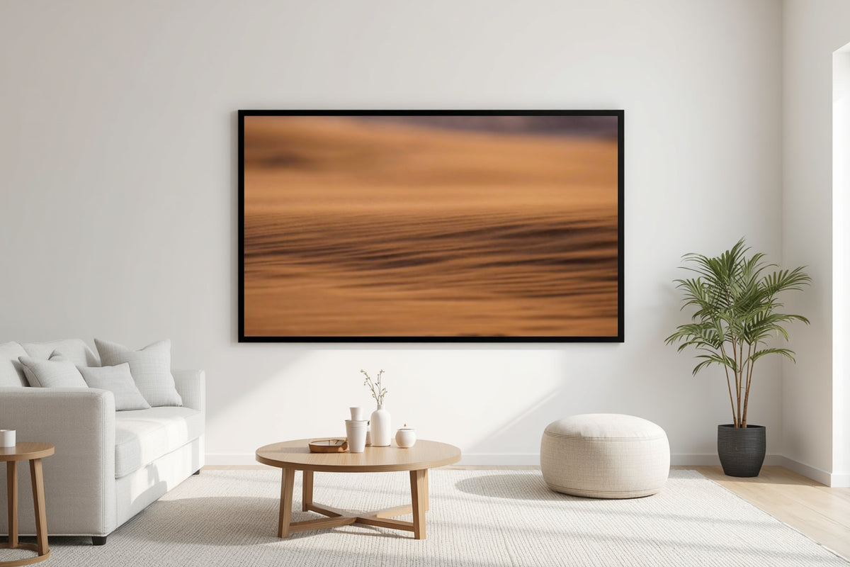A large framed abstract landscape print featuring warm orange and brown tones, displayed on a white wall in a modern, sunlit living room with a sofa, coffee table, and potted plant.