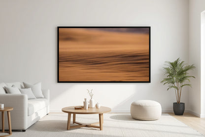 A large framed abstract landscape print featuring warm orange and brown tones, displayed on a white wall in a modern, sunlit living room with a sofa, coffee table, and potted plant.