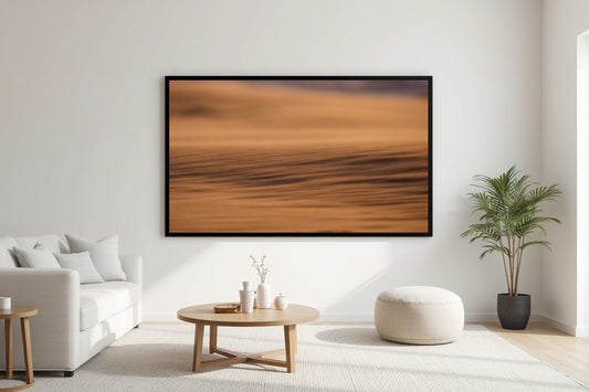A large framed abstract landscape print featuring warm orange and brown tones, displayed on a white wall in a modern, sunlit living room with a sofa, coffee table, and potted plant.