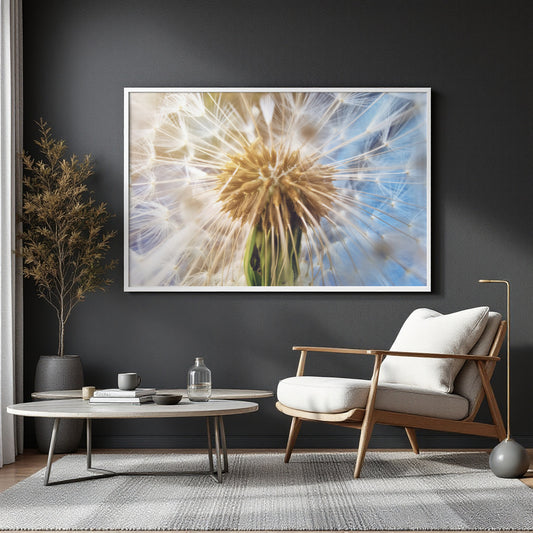 A large framed wall art print depicting a close-up dandelion in soft blue and cream tones, displayed above a modern armchair and round coffee table in a styled living room.