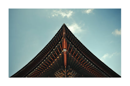 A low-angle photograph of the pointed roof corner of a traditional East Asian temple, showcasing dark tiled roofing and wooden beams against a blue sky with light clouds.