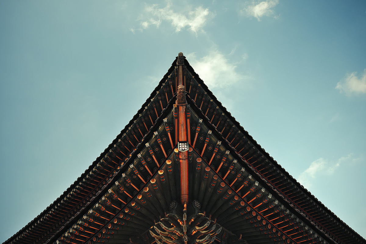 A low-angle view looking up at the sharp, symmetrical corner of a traditional East Asian temple roof against a light blue sky with soft white clouds.