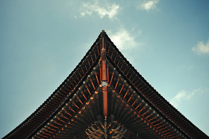 A low-angle view looking up at the sharp, symmetrical corner of a traditional East Asian temple roof against a light blue sky with soft white clouds.