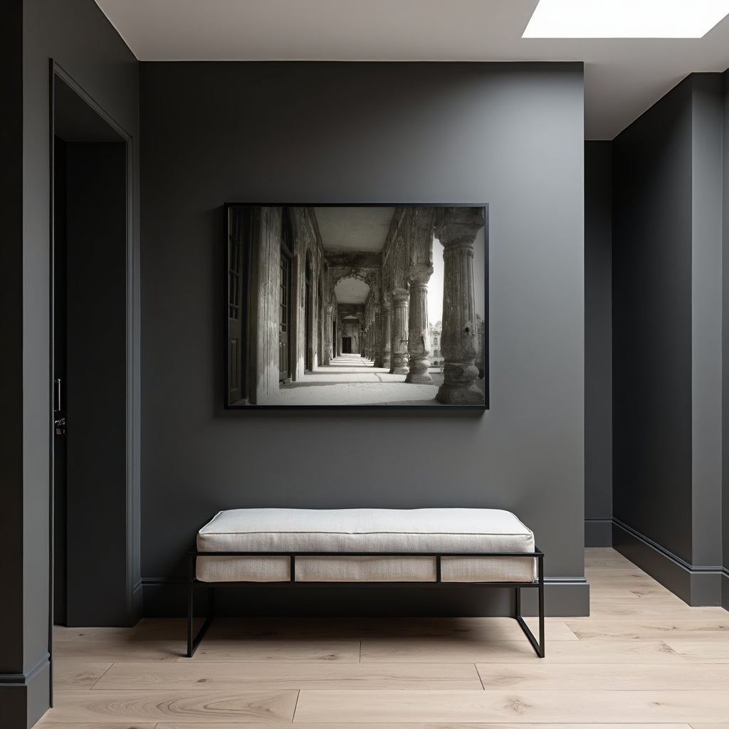 A black-framed black and white art print of an ancient stone corridor hangs on a dark gray wall above a light beige upholstered bench with black metal legs in a modern, minimalist room.