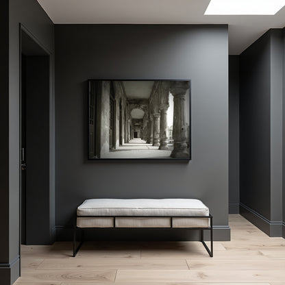 A black-framed black and white art print of an ancient stone corridor hangs on a dark gray wall above a light beige upholstered bench with black metal legs in a modern, minimalist room.
