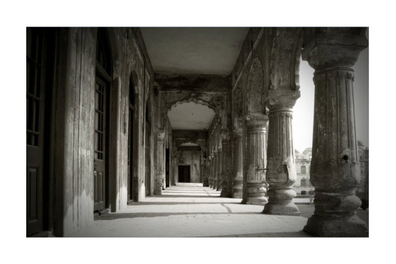Black and white architectural photograph of a historic corridor with large stone columns and arches, presented as a square print with a white border.