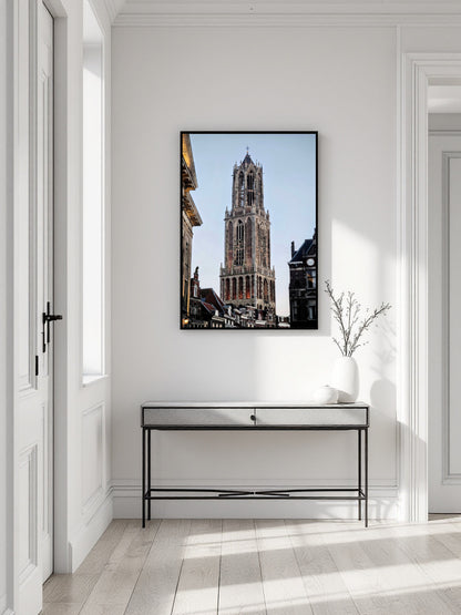 A framed vertical art print of the Belfry of Brussels under a clear blue sky, displayed on a white wall above a modern console table in a bright, minimalist hallway.
