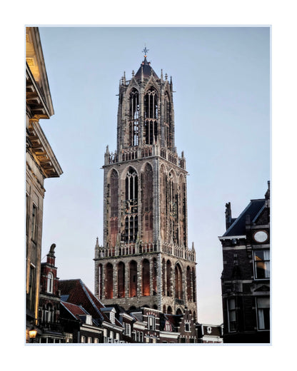 A vertical architectural photograph of the historic Belfry Tower in The Hague, Netherlands, framed by neighboring buildings against a pale blue sky.