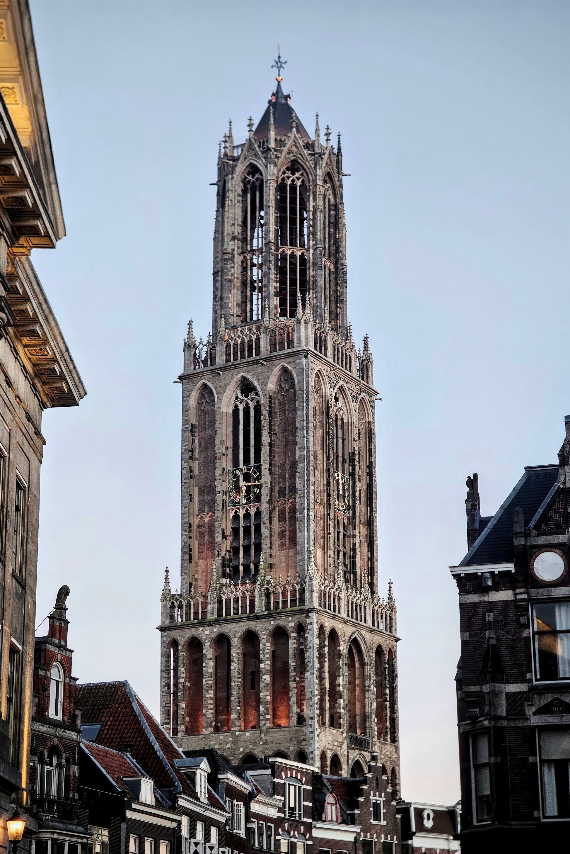 The tall stone tower of a historic church in a European city, framed by buildings on both sides against a pale blue sky.
