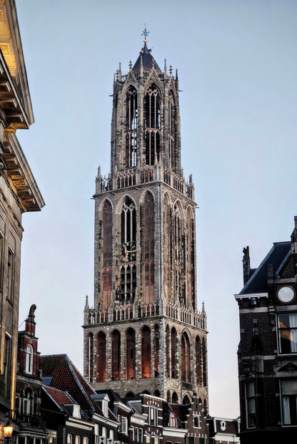 The tall stone tower of a historic church in a European city, framed by buildings on both sides against a pale blue sky.