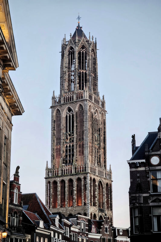 The tall stone tower of a historic church in a European city, framed by buildings on both sides against a pale blue sky.