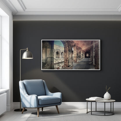 A framed horizontal art print depicting an ancient Roman-style coliseum interior, displayed on a dark charcoal wall in a modern living room setting with a blue armchair, floor lamp, and side table.