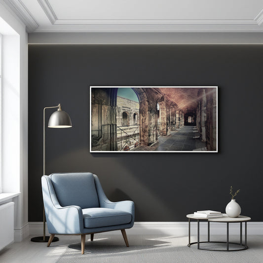 A framed horizontal art print depicting an ancient Roman-style coliseum interior, displayed on a dark charcoal wall in a modern living room setting with a blue armchair, floor lamp, and side table.