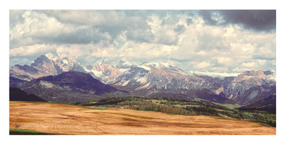 The jagged Dolomites rise beyond golden alpine fields beneath a cloud-filled sky in a photo mount
