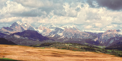 The jagged Dolomites rise beyond golden alpine fields beneath a cloud-filled sky