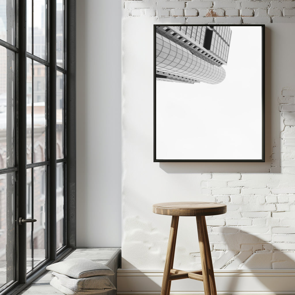 A black-framed vertical wall art print featuring a black and white architectural photograph of a curved modern building, displayed on a white brick wall above a wooden stool in a bright loft-style room.