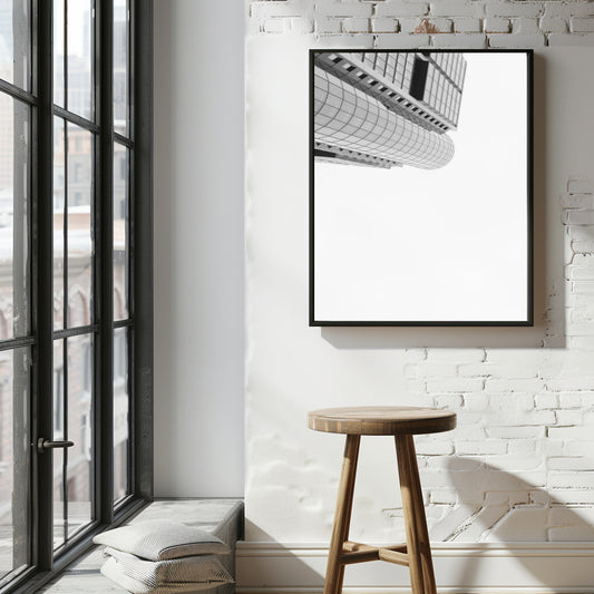 A black-framed vertical wall art print featuring a black and white architectural photograph of a curved modern building, displayed on a white brick wall above a wooden stool in a bright loft-style room.