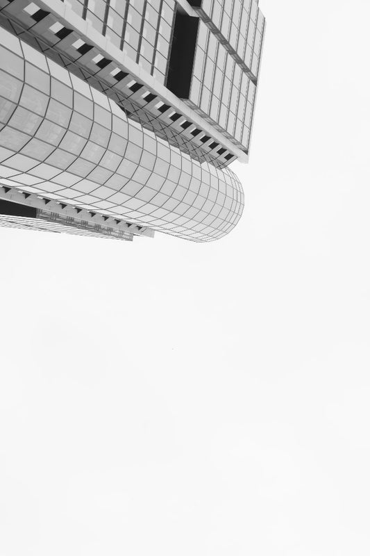 Black and white architectural photograph of a modern building with a curved glass facade, shown from a low angle against a bright white sky.