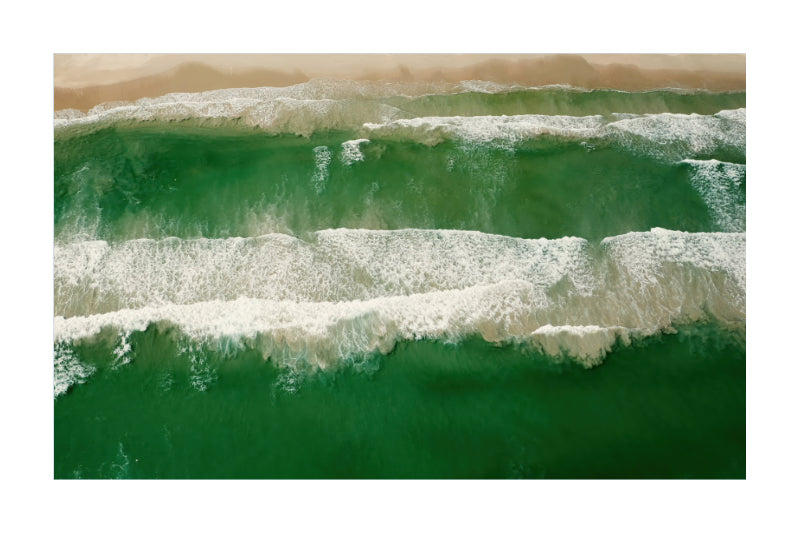 An aerial photograph of green ocean waves with white foam rolling onto a sandy beach, presented as a square art print with a white border.