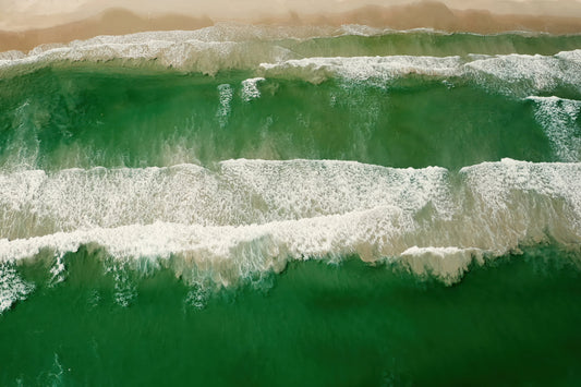 Aerial view of green ocean waves with white foam rolling onto a sandy beach.