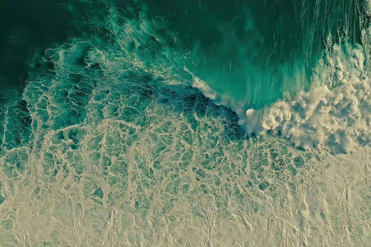 Aerial view of turquoise ocean waves with white foam crashing onto a pale sandy shore.