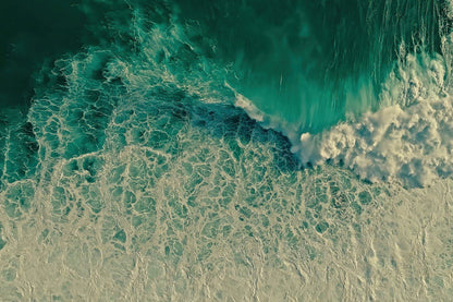 Aerial view of turquoise ocean waves with white foam crashing onto a pale sandy shore.