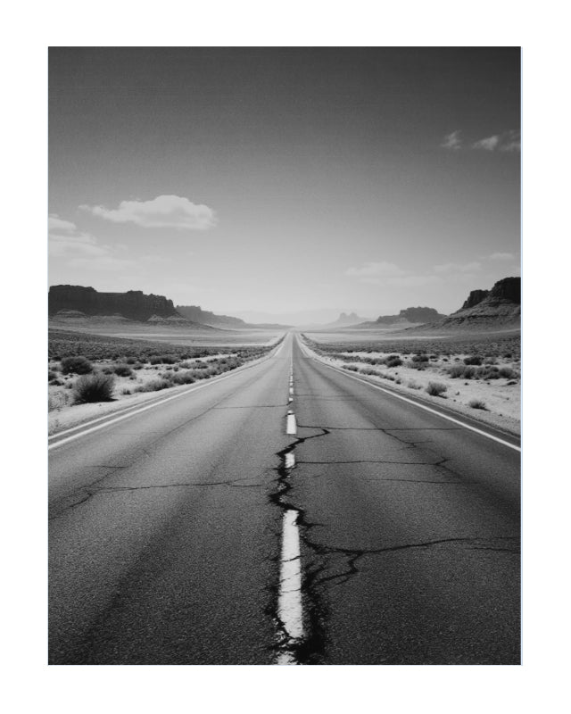 Black and white landscape photograph of a straight cracked asphalt road leading toward distant desert rock formations under a cloudy sky.