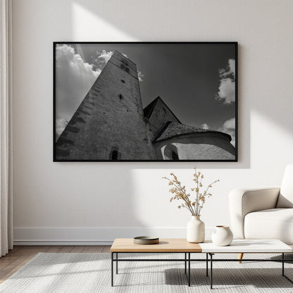 A large black and white framed wall art print depicting a stone tower and building facade under a cloudy sky, displayed in a modern living room setting above a low wooden coffee table and next to a white armchair.