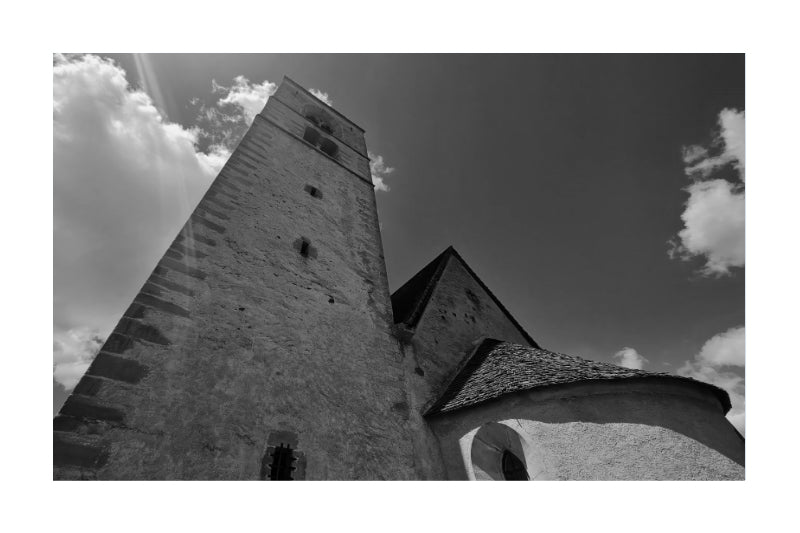 Black and white architectural photograph of a stone church tower and adjacent building under a cloudy sky, presented as wall art with a white border.