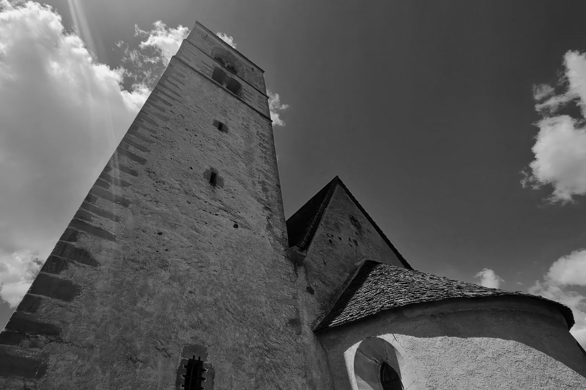 Black and white architectural photograph of a historic stone church tower and apse against a dramatic cloudy sky.