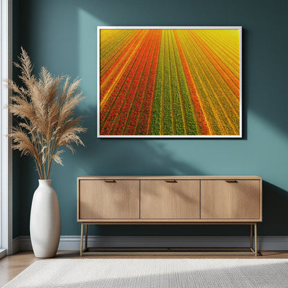 A framed art print depicting an aerial view of a vibrant field with diagonal rows of red, orange, yellow, and green crops, displayed on a dark teal wall above a light wood sideboard with a white vase of pampas grass.