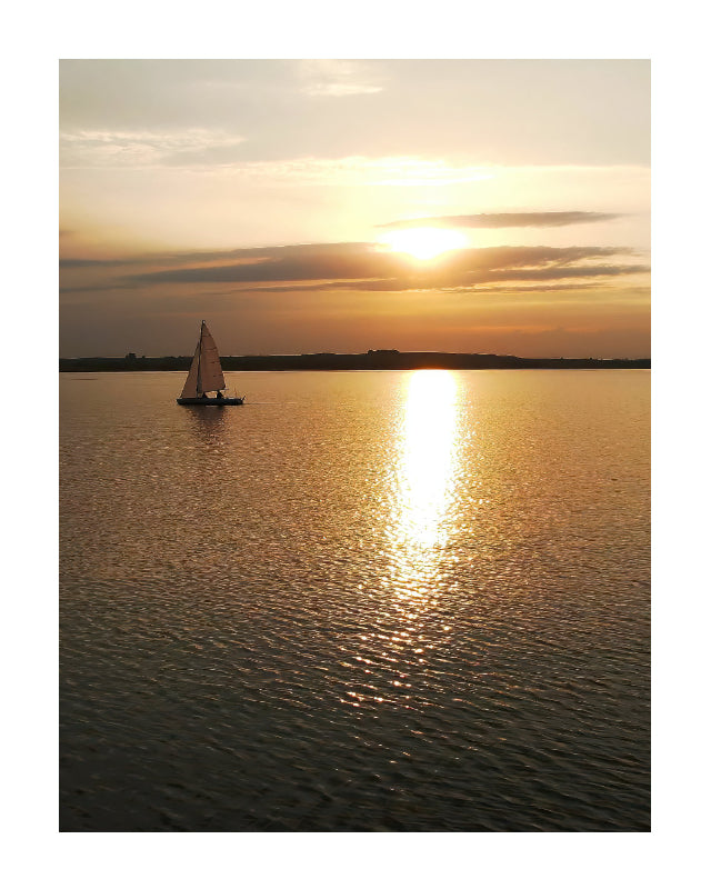 A sailboat on calm water at sunset, with golden sunlight reflecting across the surface and a dark shoreline in the distance.
