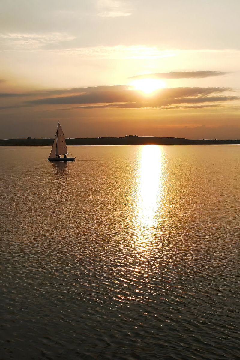 A sailboat floats on calm water at sunset, with the sun low over the horizon and bright golden light reflecting across the water surface.