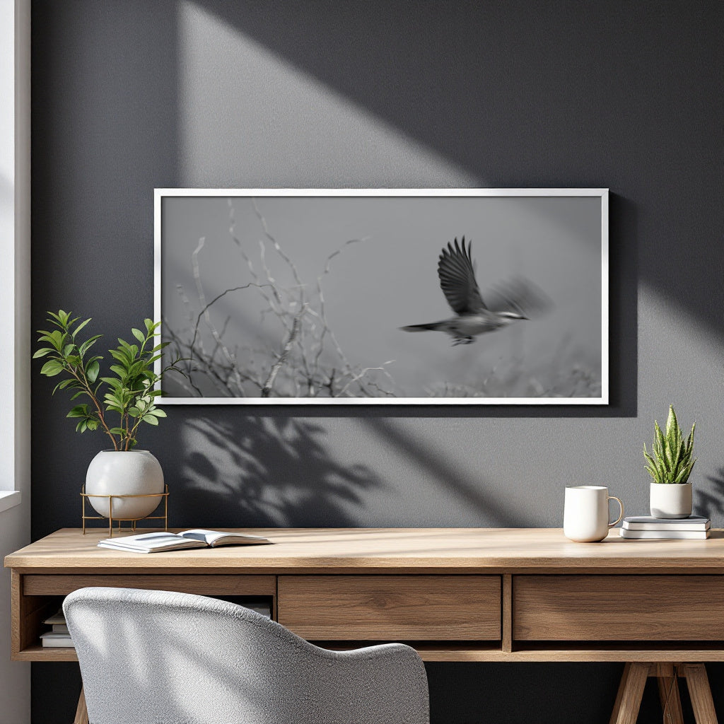 A framed black and white art print of a bird in flight hanging on a dark gray wall above a wooden desk with plants and a mug.