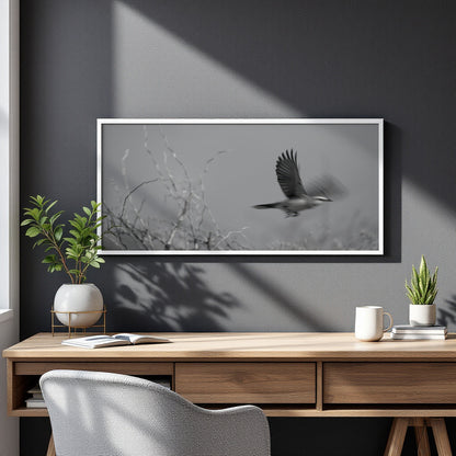 A framed black and white art print of a bird in flight hanging on a dark gray wall above a wooden desk with plants and a mug.