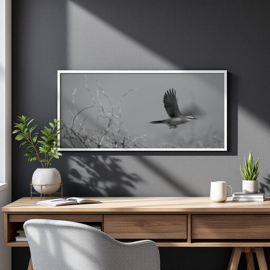 A framed black and white art print of a bird in flight hanging on a dark gray wall above a wooden desk with plants and a mug.