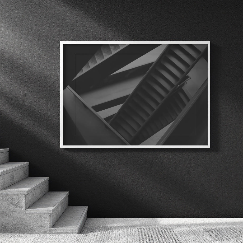 A framed black and white architectural photograph of a spiral staircase, displayed on a dark wall above a staircase in a modern interior setting.