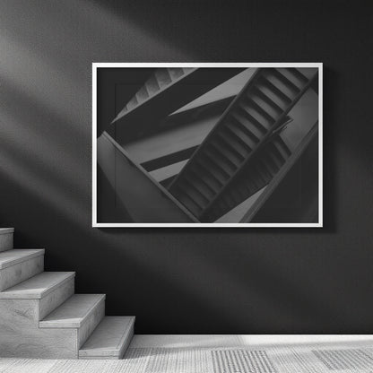 A framed black and white architectural photograph of a spiral staircase, displayed on a dark wall above a staircase in a modern interior setting.